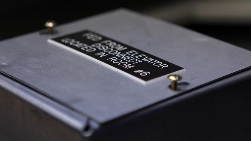 Engraved black nameplate on electrical enclosure — From Elevator Disconnect Located in Room #6