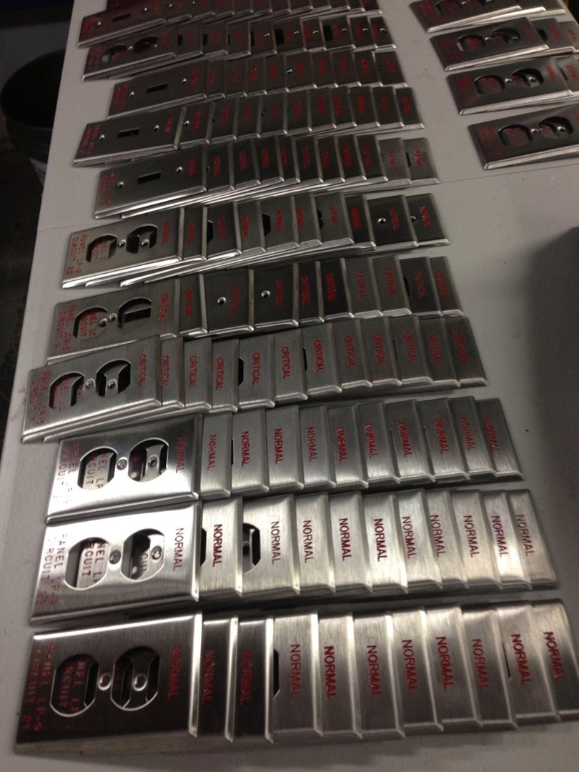 Large production run of engraved stainless steel outlet plates labeled NORMAL and CRITICAL